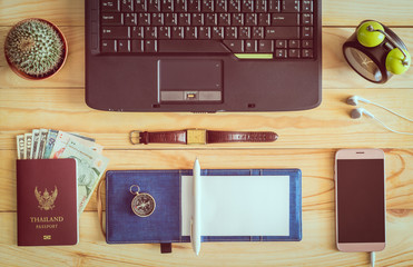 Top view of laptop, notebook, pen, passport, money, mobile phone, earphones, wristwatch, cactus, clock, and compass on wooden table.