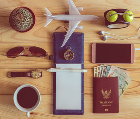 Top view of notebook, pen, sunglasses, coffee cup, passport, money, mobile phone, earphones, wristwatch, cactus, clock, airplane and compass on wooden table.