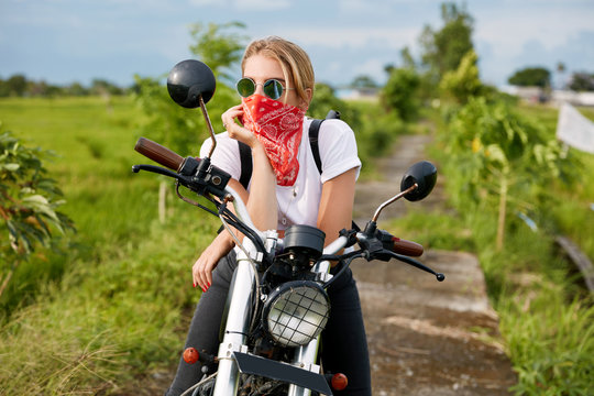 Thoughtful female biker wears shades and bandana, sits on motorbike, recreats on field, looks into distance. Hipster girl feels relaxed, carries bag, admires beautiful. People and transport concept