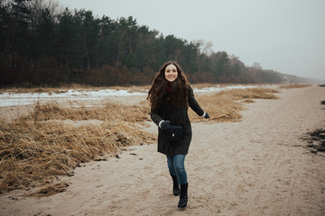Young girl in the green long jacket and blue jeans runs on the beach with black  seaweed. Jurmala Latvia