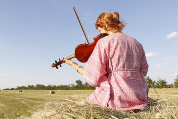 Young violinist seating on the sheaf in the wheat field and playing violin,  country side freedom...