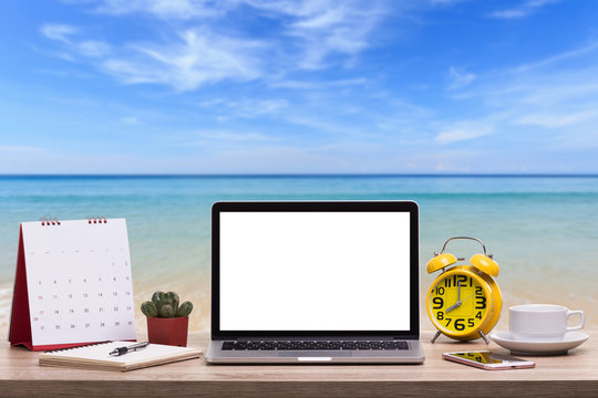 Modern laptop computer, Coffee cup, alarm clock, notebook and calendar on wooden table and view of tropical beach. Saved with clipping path. Work and holiday concept