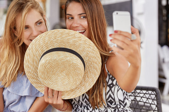 Horizontal shot of attractive beautiful women hide behind straw hat, make selfie with selphone, share their photos in social networks online. Delighted lesbian couple make photo of themselves
