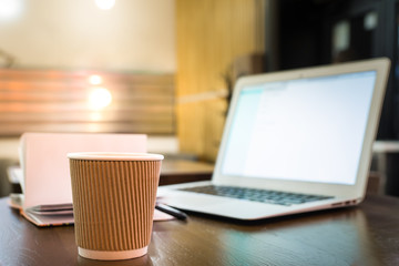 Coffee cup and laptop on the table in cafe. Portable computer, note pad and paper cup in night indoor background