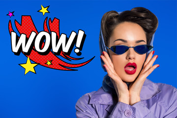 Fototapeta na wymiar portrait of stylish woman in retro clothing and sunglasses with comic style wow sign isolated on blue
