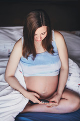 Young attractive pregnant woman is sitting on the bed with a mobile phone in hands. Beautiful pregnant woman in a rolled up tank top with a bare belly sitting on the bed and holding a cell phone.