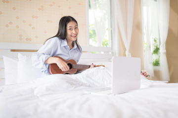 Beautiful girl practice playing ukulele on bed in the happy morning sunshine.Learn to read music chords from online computer laptop.