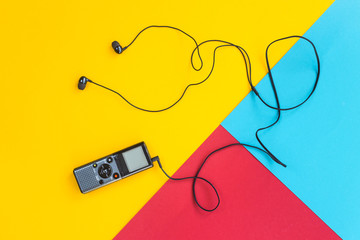 Black voice recorder with headphones on a combined yellow, blue and red background. Journalism concept. Equipment for work, interview and voice recording. Top view. Flat lay