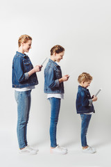 Fototapeta premium smiling mother and daughters in similar denim clothing standing in row and using tablets isolated on grey