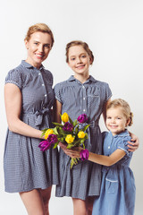 portrait of smiling mother and daughters in similar dresses with bouquet of tulips isolated on grey