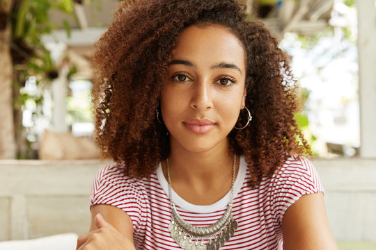 Close up shot of adorable dark skinned females with confident look, has curly hair, looks directy into camera, ponders about something. African American woman has rest against cafe interior.