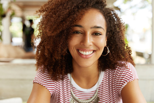 Headshot of beautiful dark skinned female with happy expression has Afro hairstyle, demonstrates perfect white even teeth, has pleased smile. Stylish young African American woman rests indoor