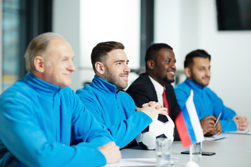 Happy young footballer in sportswear and his colleagues listening to speaker at conference