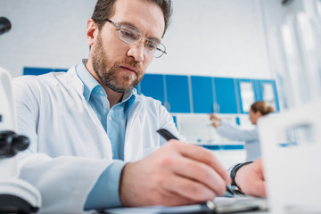 selective focus of scientist in white coat and eyeglasses making notes in notepad at workplace in laboratory