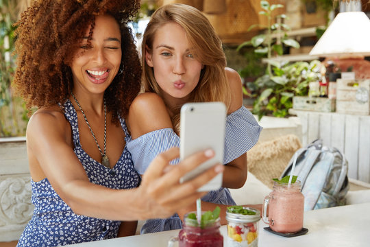 Positive female best friends pose for selfie, use modern cell phone, sit at restaurant, have funny expressions, share photos in social networks, connected to wireless internet. Friendship concept