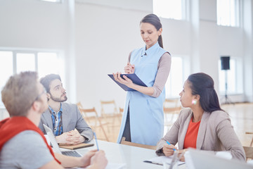 Obraz premium Young confident coach standing by desk and making notes in document or plan while three young managers listening to her
