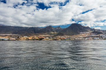 Naklejka premium Vibrant scenery and deep-blue waters of the Tenerife west coastline as seen from a yacht. The dormant Teide volcano can be seen in the background. Many tourism resorts overlook the coastline.