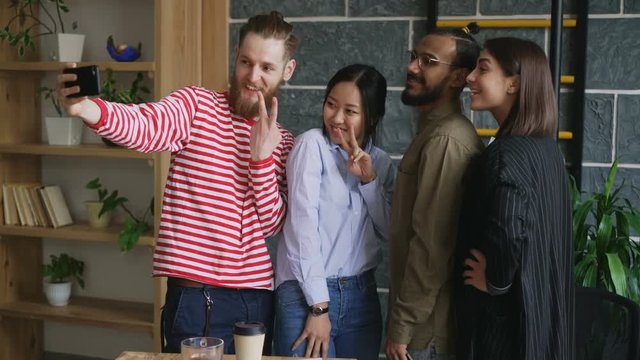 Happy multi-ethnic team taking selfie in modern loft office. Hipster man holding smartphone take self portrait with his colleagues