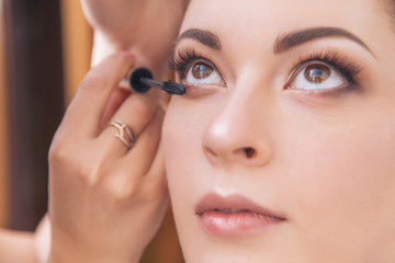 Professional make up artist apply mascara to a young caucasian woman on her date and wedding photosession. Indoors, studio. Casual outfit. Backstage of a process