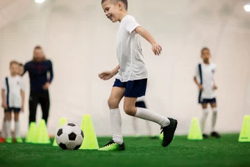 Gordijnen Happy boy in uniform kicking the ball while running around cones during training © pressmaster