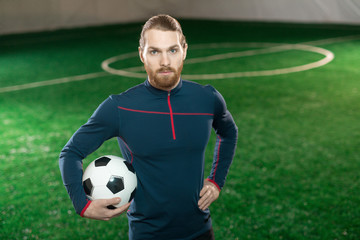 Young confident football trainer in sportswear looking at camera while standing on pitch