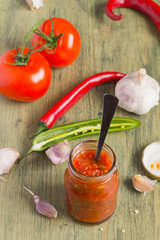 Georgian Adjika sauce in a small glass jar, next to the ingredients for its preparation on an old wooden table. 
