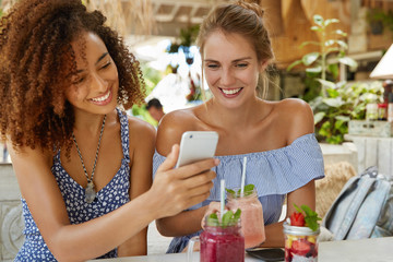 Photo of glad female with dark skin and Afro hairstyle spends free time together with girlfriend, have homosexual relationships, use modern cell phone for making selfie, enjoy tasty cocktails