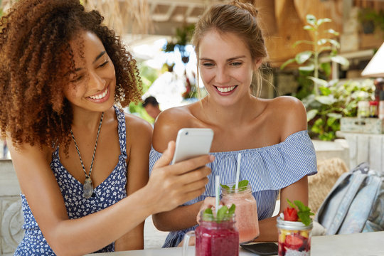 Photo of glad female with dark skin and Afro hairstyle spends free time together with girlfriend, have homosexual relationships, use modern cell phone for making selfie, enjoy tasty cocktails