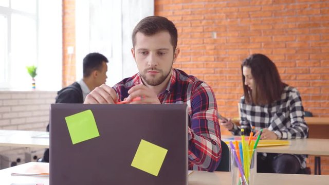 A young attractive man sits in front of a computer and twists a pencil in his hand. The concentrated man looks closely at the monitor. Shooting in the office. in the background, the extras from the