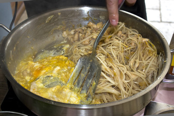 A man's hand fry rice noodles with chicken and eggs in a large professional pan