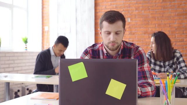 A young attractive man sits in front of a computer and twists a pencil in his hand. The concentrated man looks closely at the monitor. Shooting in the office. in the background, the extras from the