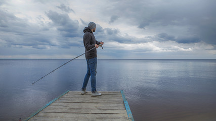 Young handsome brutal caucasian man in casual outfit fishing on a lake as a hobby, trying to catch carp on spinning rod alone. Summer weather, sunset, natural landscapes. HDR toned image