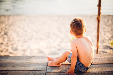 The theme is a child and summer beach vacation. A small boy sits with his back on a wooden pier and admires the view of the sandy beach and the pond, the river. With bare legs in blue denim shorts