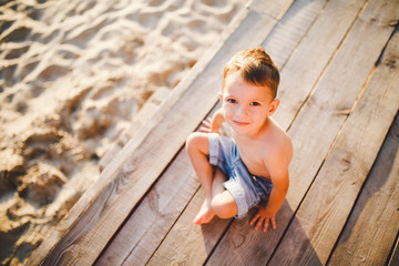 The theme is a child and summer beach vacation. A small Caucasian boy sits sideways on a wooden pier and looks at the camera on a sandy beach and a pond, a river. With bare legs in blue denim shorts