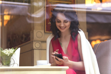 Fototapeta na wymiar Beautiful brunette caucasian business woman sitting in cafe on a coffee break in red casual but a bit classic dress and white cardigan, smile, talking on the phone. Copy space. Evening, sunset light
