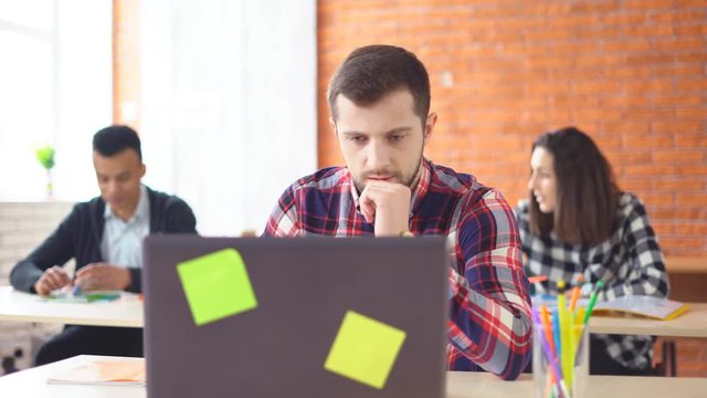 young attractive man sitting in front of computer and typing on laptop. The concentrated man looks closely at the monitor. Shooting in the office. in the background, the extras from the actors. 4k