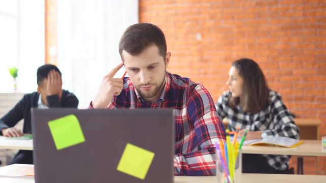 young attractive man sitting in front of computer and typing on laptop. The concentrated man looks closely at the monitor. Shooting in the office. in the background, the extras from the actors. 4k