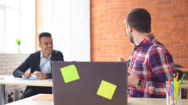 young attractive man sits in front of a computer and communicates with colleagues. a friendly atmashere in the classroom. people are laughing. Shooting in the office. in the background, the extras