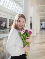 Portrait of attractive young woman with tulips is standing in light room and smiling. Happy international women's day! Celebrating 8th of March. Happy girl smiling and holding pink tulip flowers.