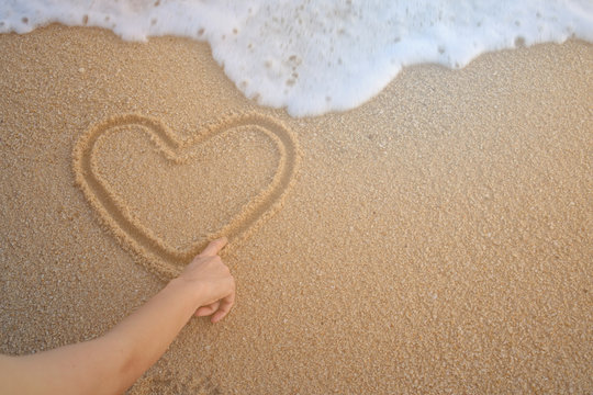 Hand of the women drawing heart shape  on the sand of a beach with wave of the sea.    