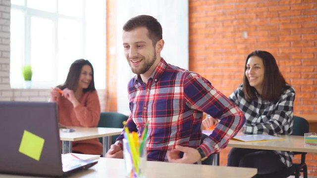 young attractive man sits in front of a computer and communicates with colleagues. a friendly atmashere in the classroom. people are laughing. Shooting in the office. in the background, the extras