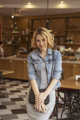 Blonde (with dark roots) caucasian woman in casual summer outfit at the cafe. Grey dress and jeans jacket. Woman got natural day makeup and curly hairstyle. She talks on her phone, working, make calls