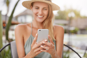 Smiling young woman with positive expression, holds smart phone while sits at terrace cafe, texts messages or surf social networks, connected to wireless internet. Summer lifestyle and rest.