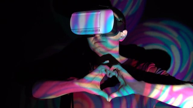 Young man in virtual reality glasses making heart shape with hands on bright spotlight background