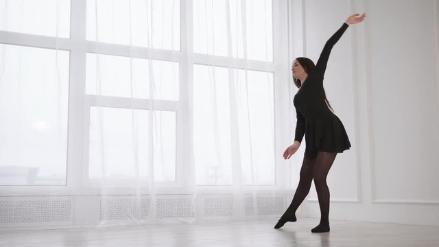 Ballerina is doing exercises for stretching back and hands. She is standing in a light hall and tiping, tensioning her hands