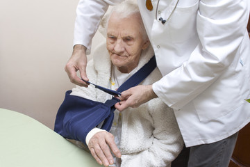 Bardzo stara siwa kobieta siedzi na krześle. Lekarz zakłada temblak na rękę siwej staruszki.