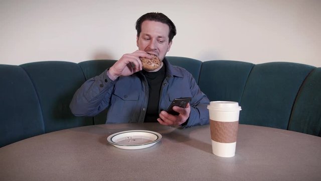 Man Scrolling Smartphone Eating Bagel at Coffee Shop