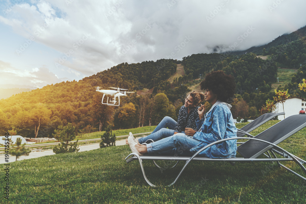 Wall mural Two charming cheerful girls of different races on deck chairs on the lane are sitting to the camera of the flying drone while it filming them; with hill ridge in the background; autumn morning