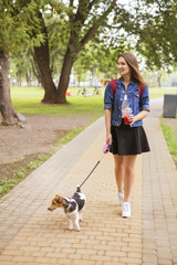 Cute beautiful slim caucasian brunette woman in casual outfit (skirt, shirt and jeans jacket) on a walk in park in summer day with her adorable jack russell dog. They full of joy, smiling and have fun