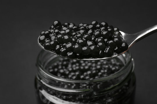 Jar and spoon with delicious black caviar on dark background, closeup